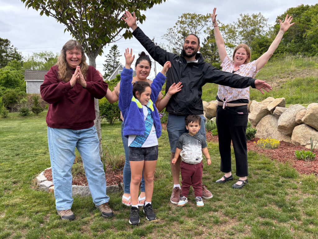 Group of smiling adults and children with arms raised, standing on a lawn in front of trees and rocks